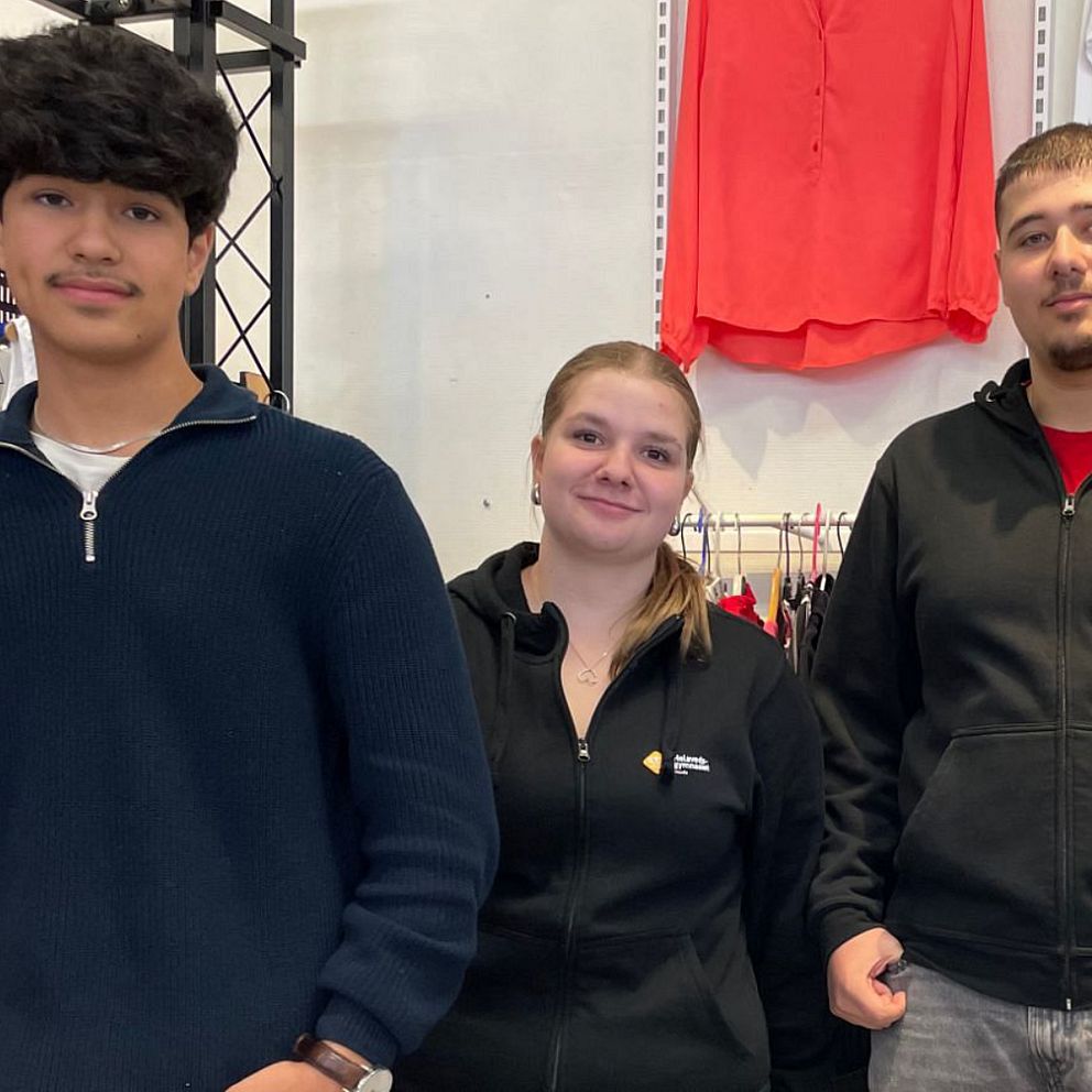 Tre personer klädda står framför en vägg med kläder. På höger sida finns en displayhylla med en rad olika plagg, inklusive tröjor och skjortor, som är organiserade på ett prydligt sätt.