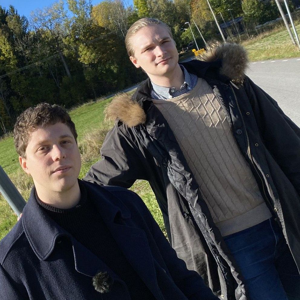 Två personer står på en grön gräsyta med träd i bakgrunden, iförda varma kläder. Till höger syns en mörk väg som sträcker sig in i natten, belyst av sporadiska ljus längs sidan.