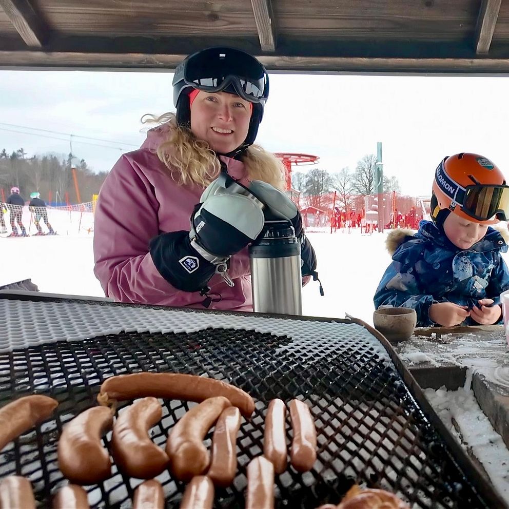 I förgrunden, korvar på grillgaller. Bakom en kvinna i rosa skiddräkt och slalomhjälm