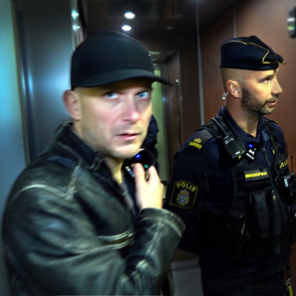 Reportern Patrik med en läderjacka står nära en polis med uniform som knackar dörr i en korridor i ett lägenhetshus.