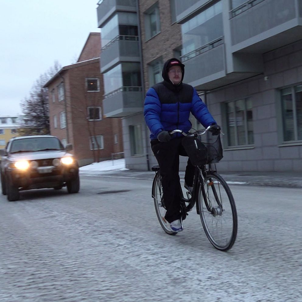 En delad bild med två sektioner. Den vänstra sektionen visar en cyklist i vinterkläder som cyklar på en snöig gata, medan en bil passerar i bakgrunden och flera byggnader syns i omgivningen. Den högra sektionen visar en närbild på Sara Keisu-Lundh (S) som pratar, med fokus på kläderna och mikrofonen.