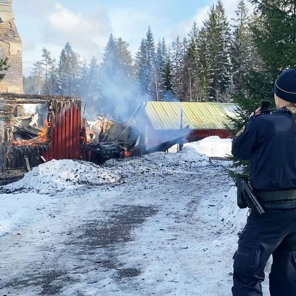 Brandman och polis vid utbränd byggnad i snöigt landskap. Rök stiger från ruinerna.