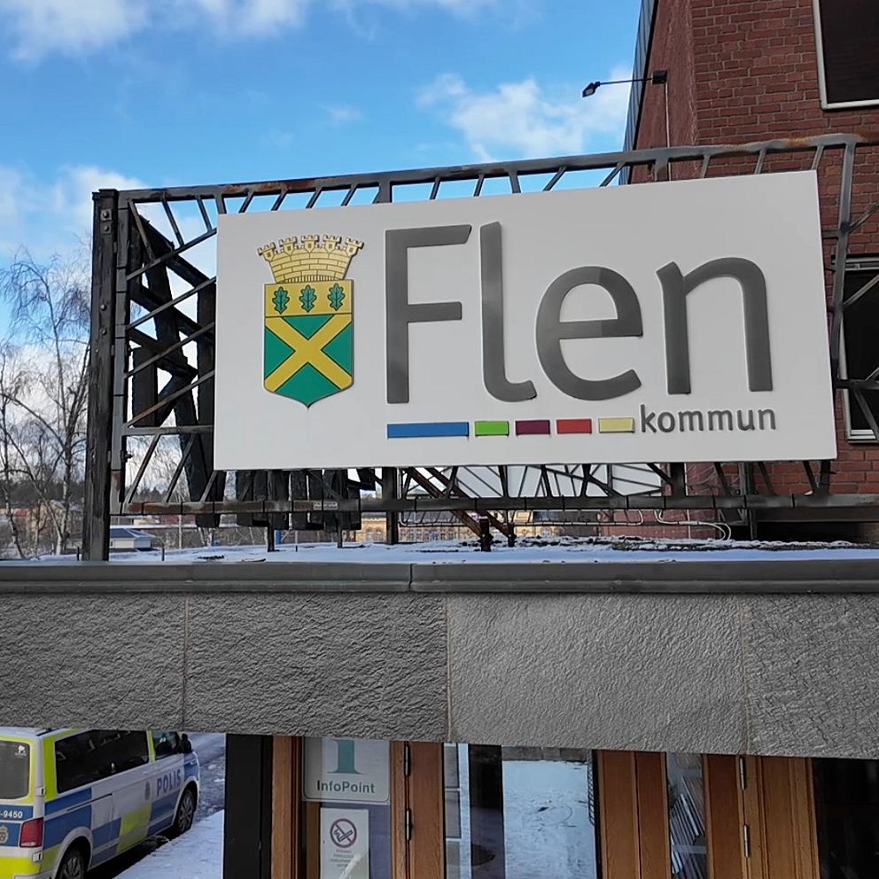 Delad bild: Till vänster syns en skylt med texten ”Flen kommun” monterad på en byggnad med en polisbil delvis synlig i bakgrunden. Till höger står en person med långt hår, klädd i en stickad tröja och bär en mikrofon.