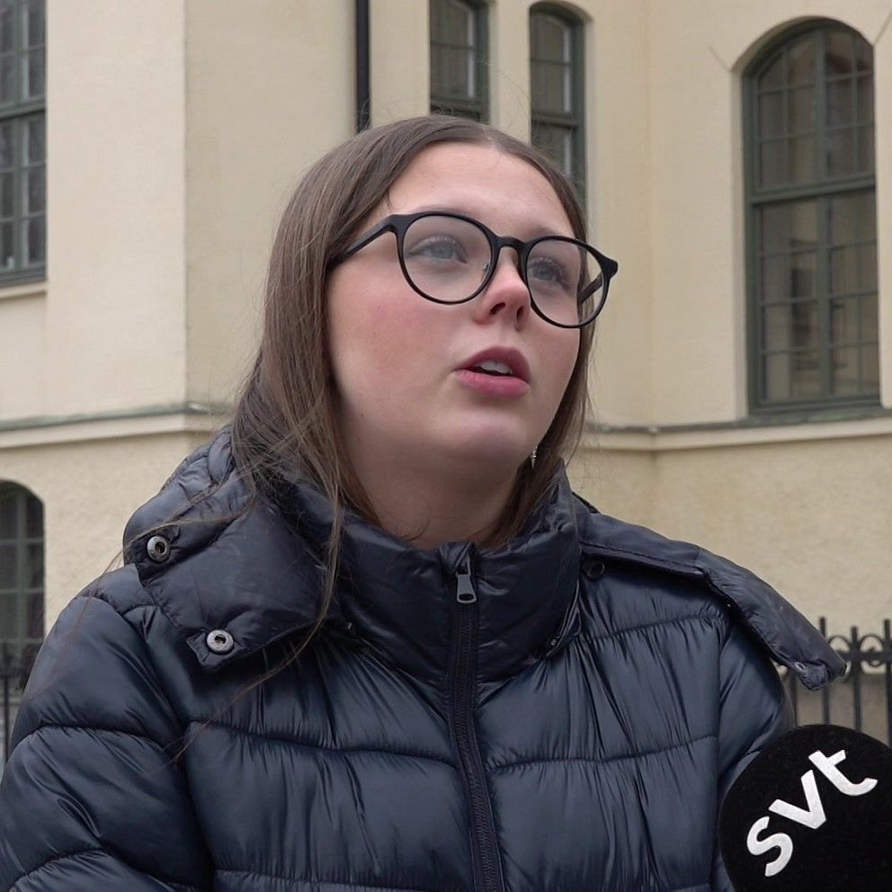 En person intervjuas utomhus av SVT framför en beige byggnad med stora fönster. Personen bär en mörk, vadderad jacka och håller en SVT-mikrofon. I bakgrunden syns ett staket och en flaggstång.
