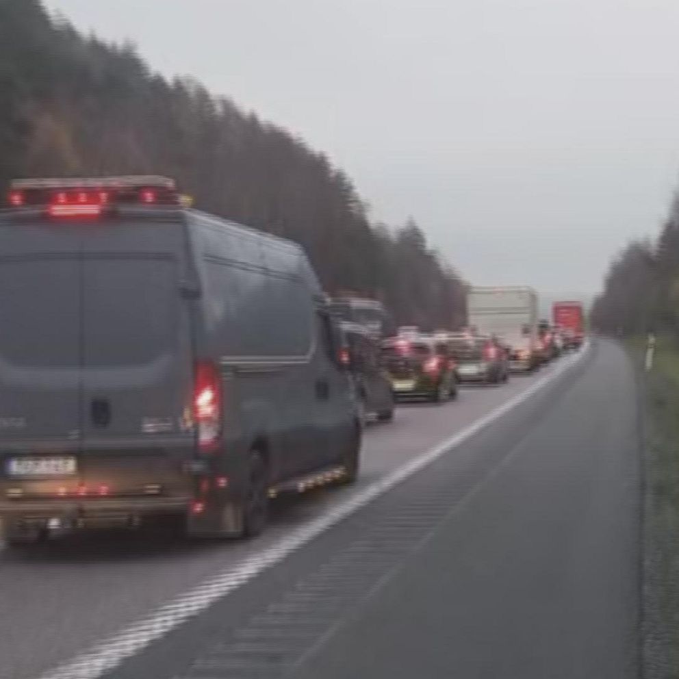 Kö av bilar på en motorväg, med en svart skåpbil i förgrunden och färre bilar längre fram. Omgivningen är dämpad med träddel och gräsmatta vid vägkanten under molniga förhållanden.