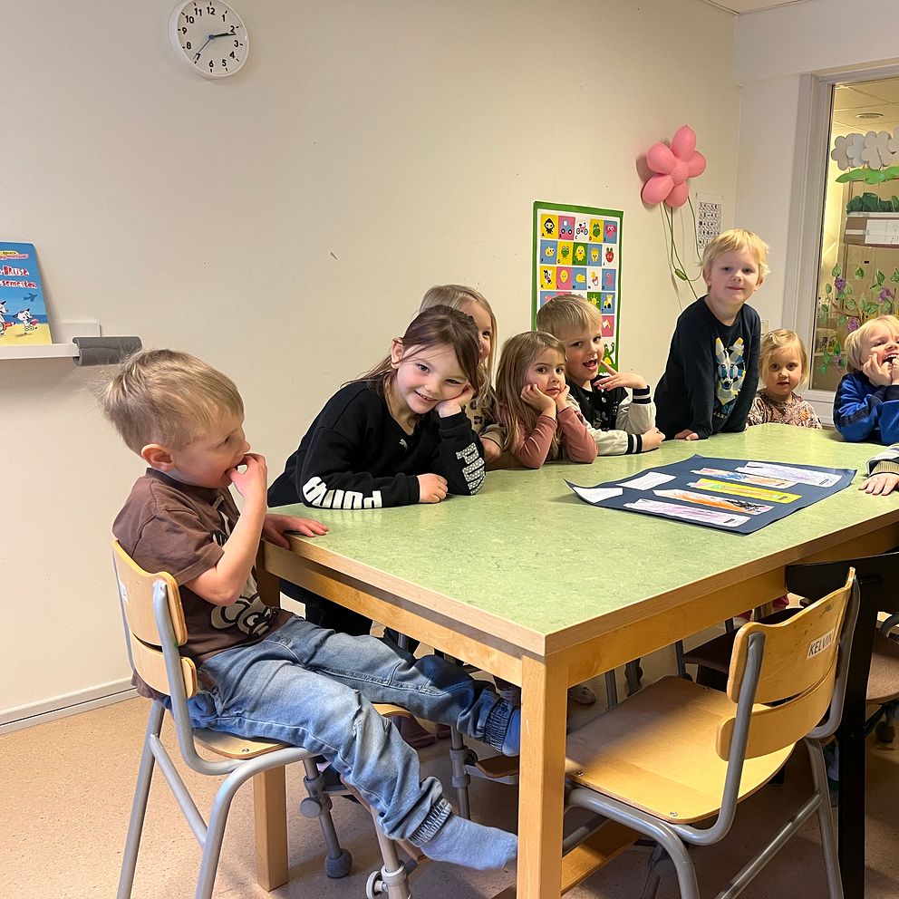 Nio förskolebarn som sitter och står runt ett bord