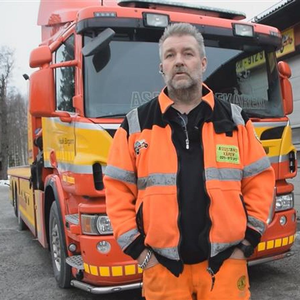 Bärgaren Peter Norén i orange reflexjacka och byxor står framför en stor, ljusorange och gul lastbil vid en verkstad. Bakgrunden visar en byggnad med garageportar och snö på marken, vilket indikerar en vintermiljö.