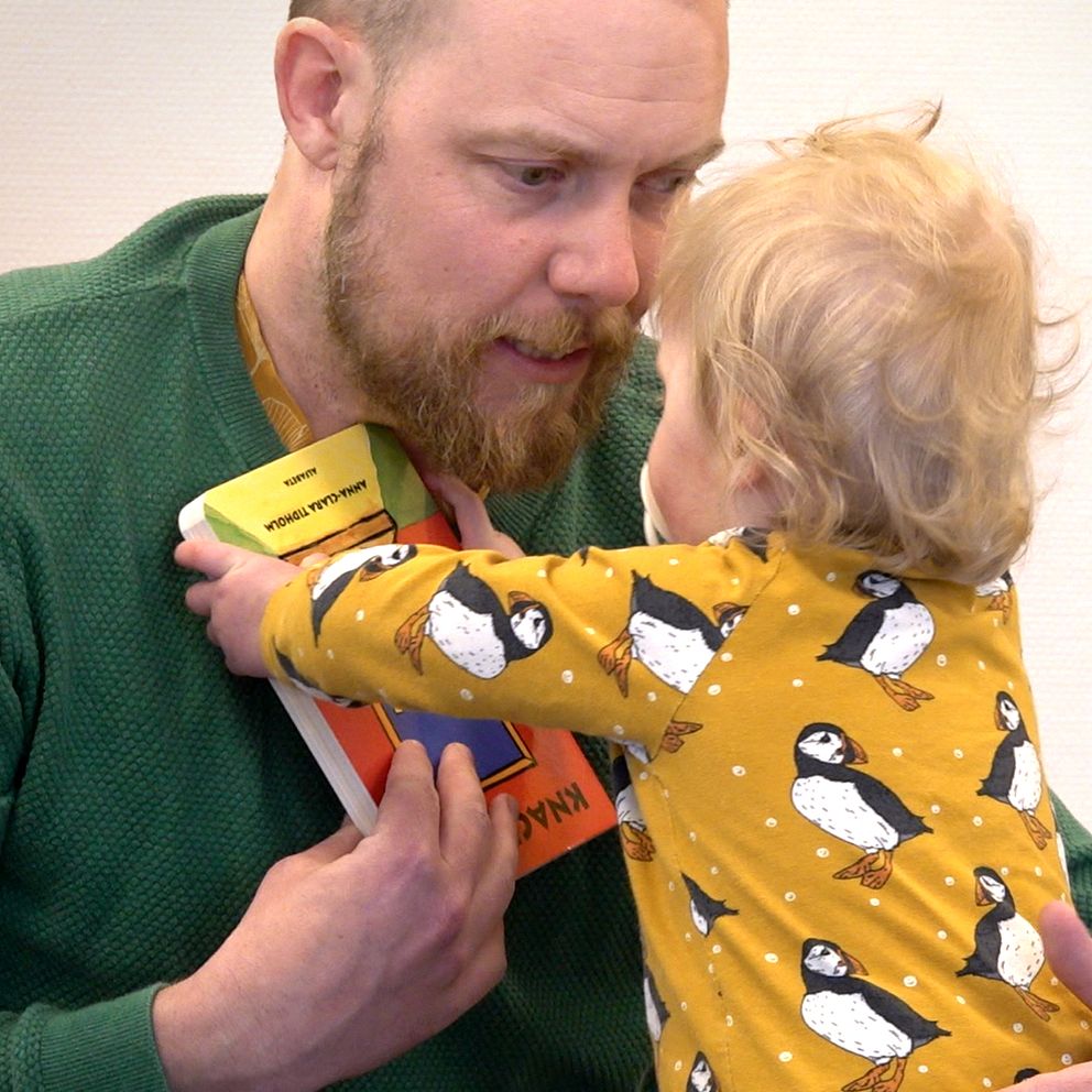 en man med ett barn i famnen som håller fram en barnbok.
