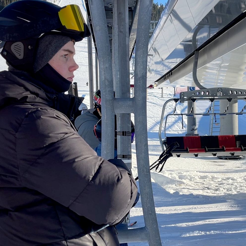 En person iförd hjälm och skidkläder står bredvid en sittlift på en snötäckt skidort, med skidliftstolar i bakgrunden.