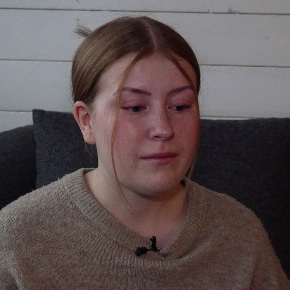 En person med brunt hår sitter i en soffa klädd i en beige tröja och har en mikrofon fäst på tröjan. Bakgrunden består av en vit träpanel och mörka soffkuddar.