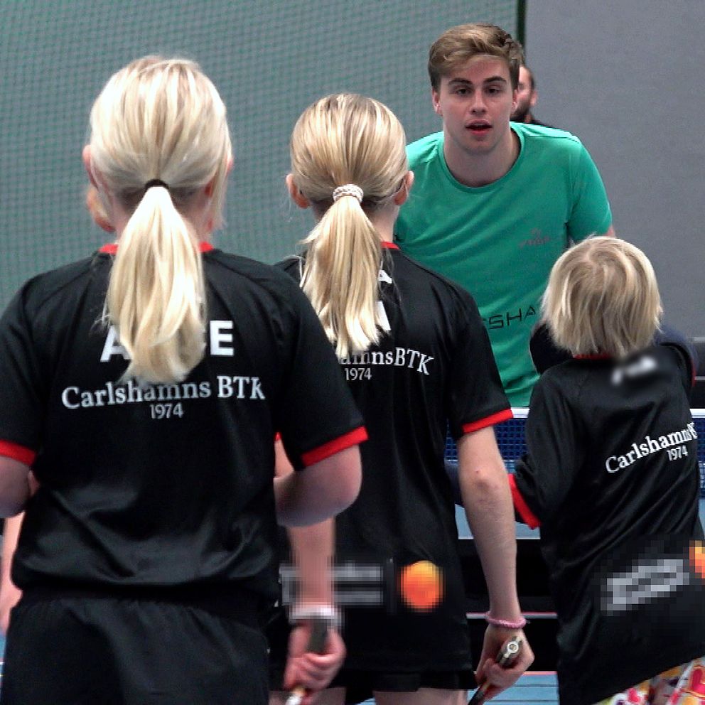 Tre unga spelare i svarta tröjor med texten ”Carlshamns BTK” står vid ett bordtennisbord och vänder sig mot Truls Möregårdh som ger instruktioner. En vuxen person syns i bakgrunden, också i träningskläder. Hallen har en sportig atmosfär med nät och annan utrustning synlig i omgivningen.