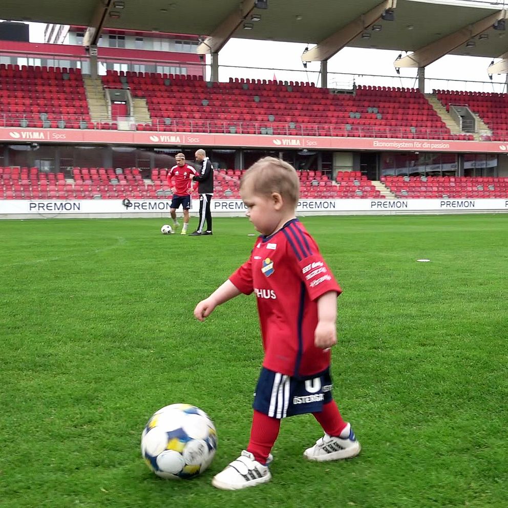 Litet barn i röd matchtröja spelar fotboll på arena, vuxen man tittar på i bakgrunden.