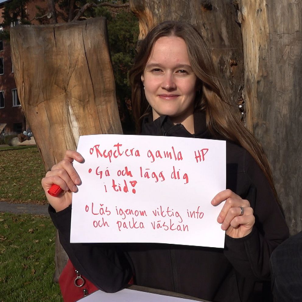 Rebecca Zauner håller upp en skylt med texten ”Repetera gamla HP” samt ”Gå och lägg dig i tid” och ”Läs igenom viktig info och packa väskan” i en utomhusmiljö med träd och en byggnad i bakgrunden.