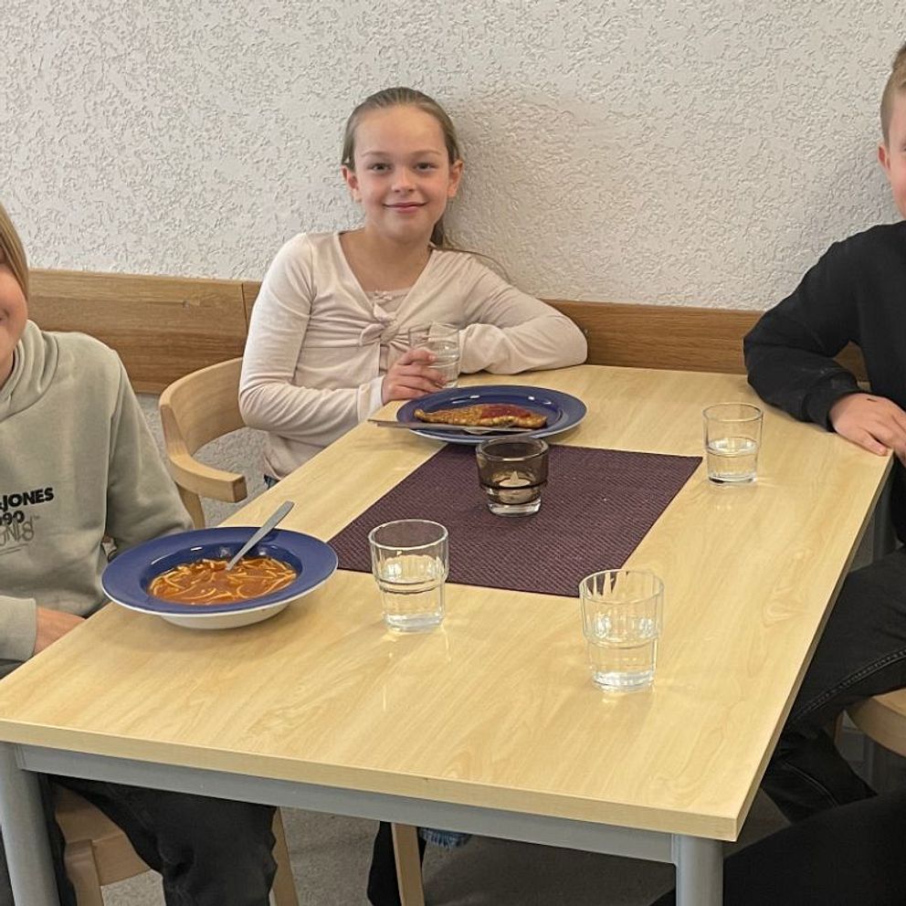 Fyra ungdomar sitter vid ett bord i en matsal, två håller tallrikar med soppa.