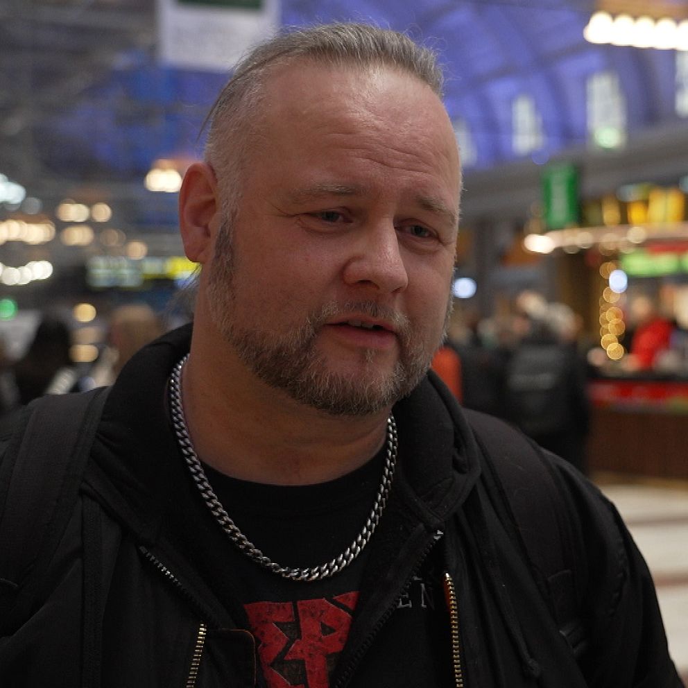 Man i svarta kläder med silverkedja står i en ljus, livlig tågstation med matställen och människor i bakgrunden.