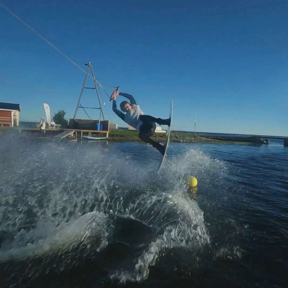 En person utför en tricksurfingmanöver på en wakeboard på wakeboardbanan i Långviken i Kalmar i en stilla vattenmiljö, omgiven av en klar blå himmel och några byggnader i bakgrunden. Vattnet stänker upp kring Oskar Sörman från Kalmar wakeboardförening, som är i luften med en surfbräda under fötterna.