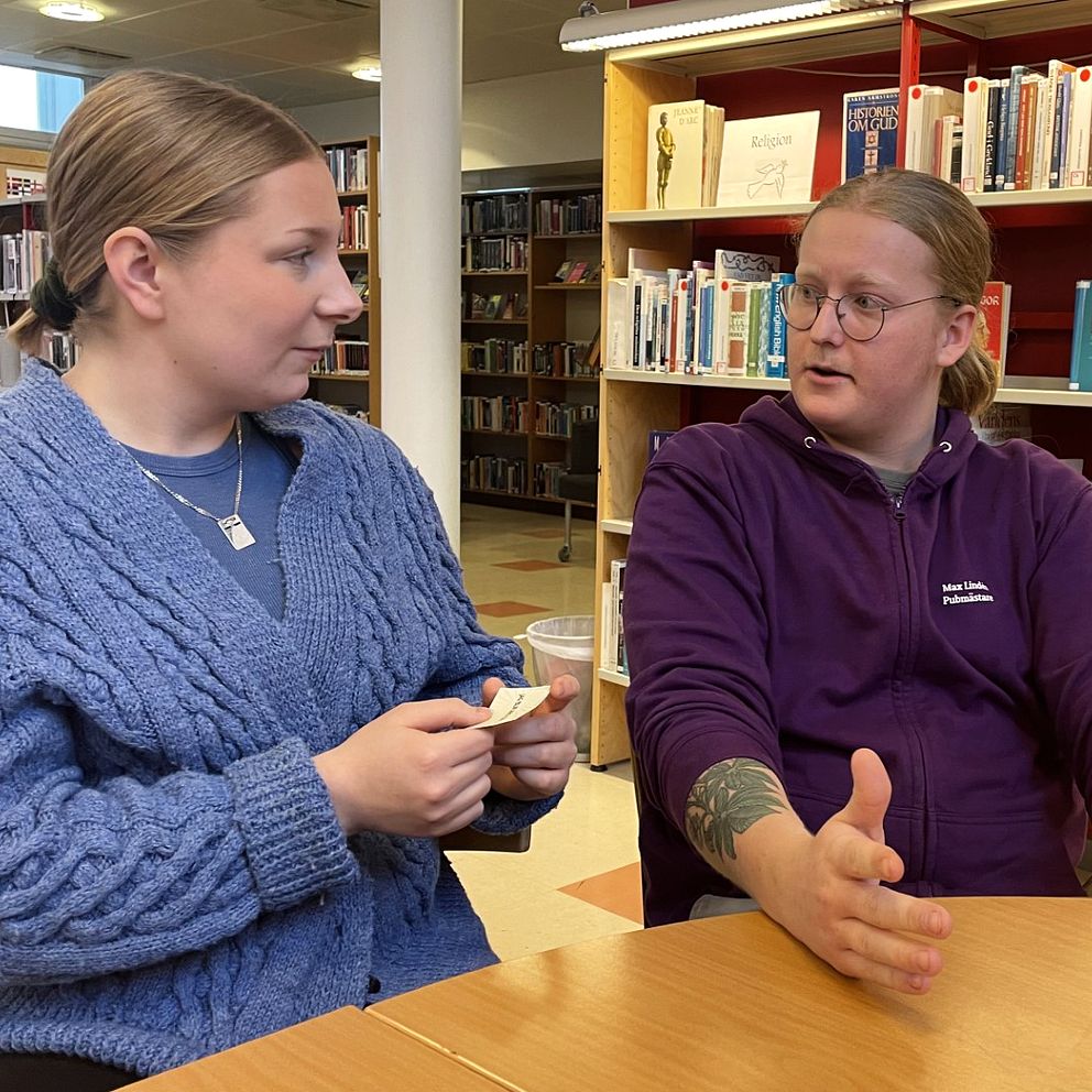 Johanna Elg och Max Lindén sitter vid ett bord i biblioteket på Olympiaskolan i Helsingborg, omgivna av bokhyllor i bakgrunden. Johanna bär en blå kofta och håller upp ett kort i handen, medan Max är klädd i en lila tröja och gestikulerar med händerna. Atmosfären är avslappnad och de  diskutera något.