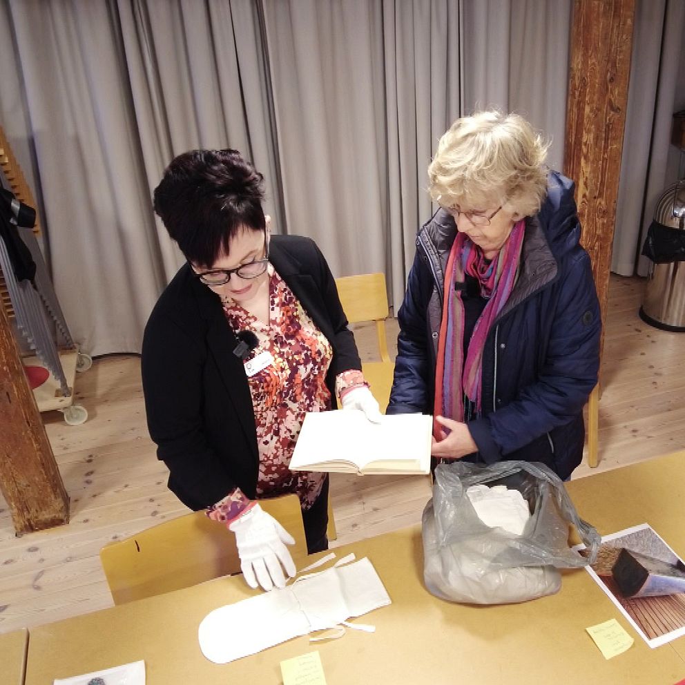 Gerd Nilsson visar en bok och en gördet till en blöja för en samlingsintendenten på Rian museum i Falkenberg. På bordet finns en grå plastpåse, en vit handske och flera föremål med klisterlappar, inklusive tidskrifter och andra småsaker.