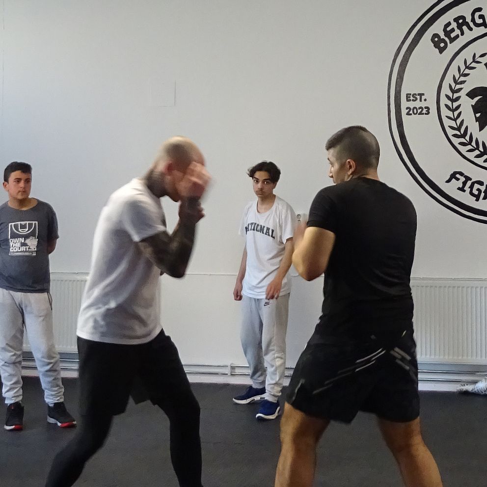 En intensiv sparringmatch fångas i aktion på Bergslagens Fight Club, där två atleter utövar sina färdigheter. I bakgrunden observerar tre personer noggrant, medan klubbens emblem – en dubbelhövdad örn och Bergslagen-flaggan – dominerar väggen