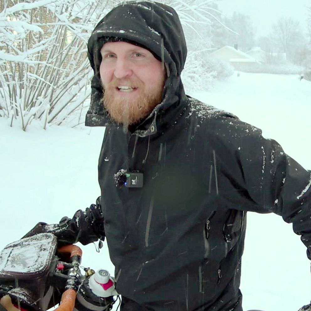 En person klädd i svart vinterjacka står i ett snöigt landskap, omgiven av snöiga träd och en väg som leder längre bort. Personen är utrustad med en cykel och har olika tillbehör kopplade till cykeln. Snön faller runt dem, vilket skapar en kall och vintrig atmosfär.
