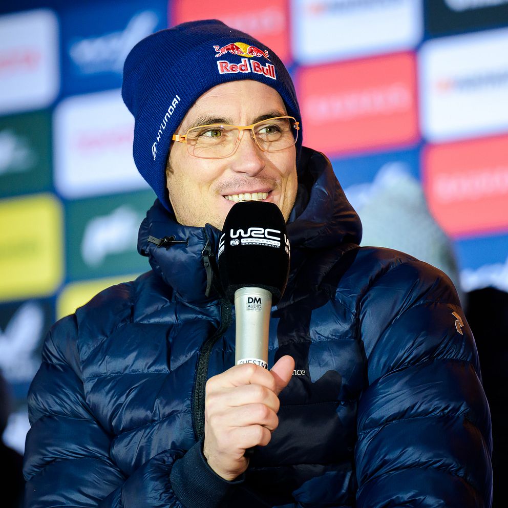 Thierry Neuville i mörk jacka håller en mikrofon framför en bakgrund med olika logotyper och färgglada rutor.