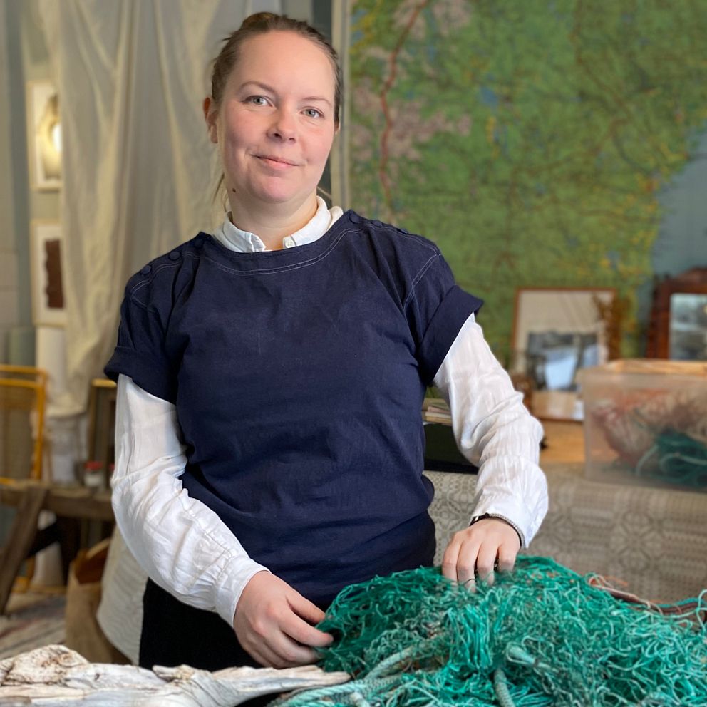 Konstnären Tove Gullikson står vid ett bord och arbetar med grönt fisknät. Bredvid står en svart och vit katt. I bakgrunden syns en karta över södra Sverige.