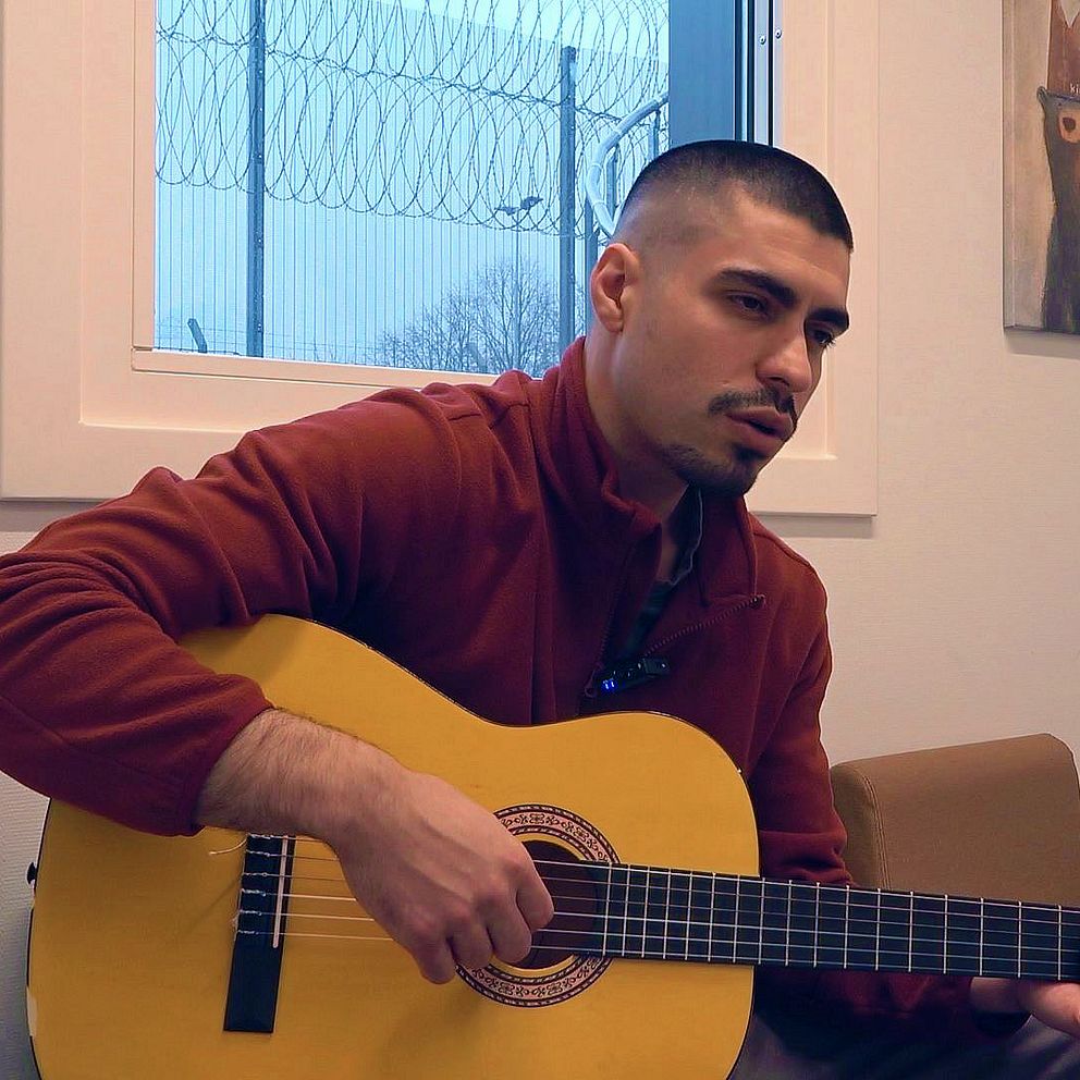 Kille spelar på akustisk gitarr. Fönster med taggtrådsstängsel utanför