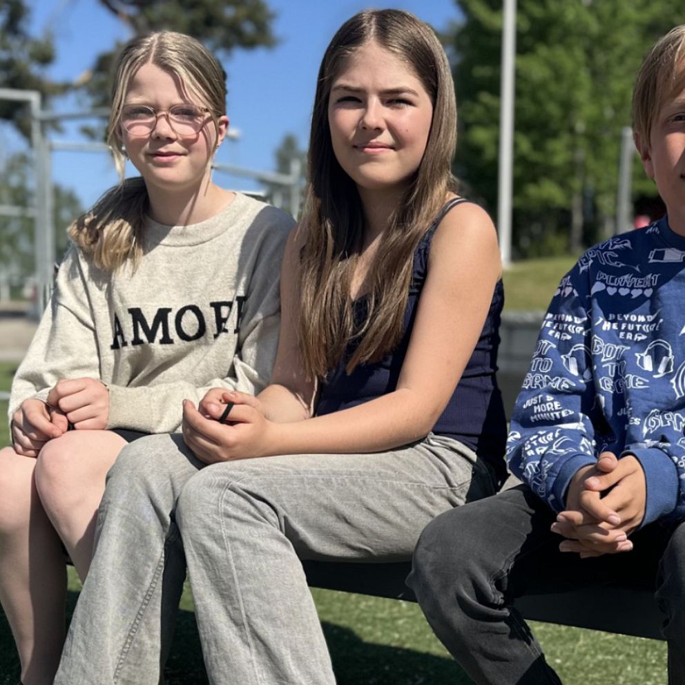 Tre elever, två tjejer och en kille som går i årskurs fem sitter på en bänk utomhus på skolgården.