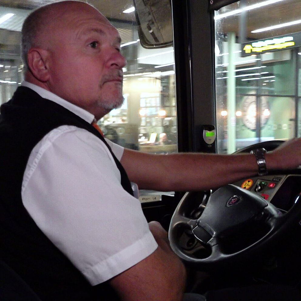 Busschauffören Suad Kaharevic sitter i en regionalbuss i Helsingborg, iklädd arbetskläder som inkluderar en väst och skjorta. Han sitter i förarsätet och ler mot kameran.