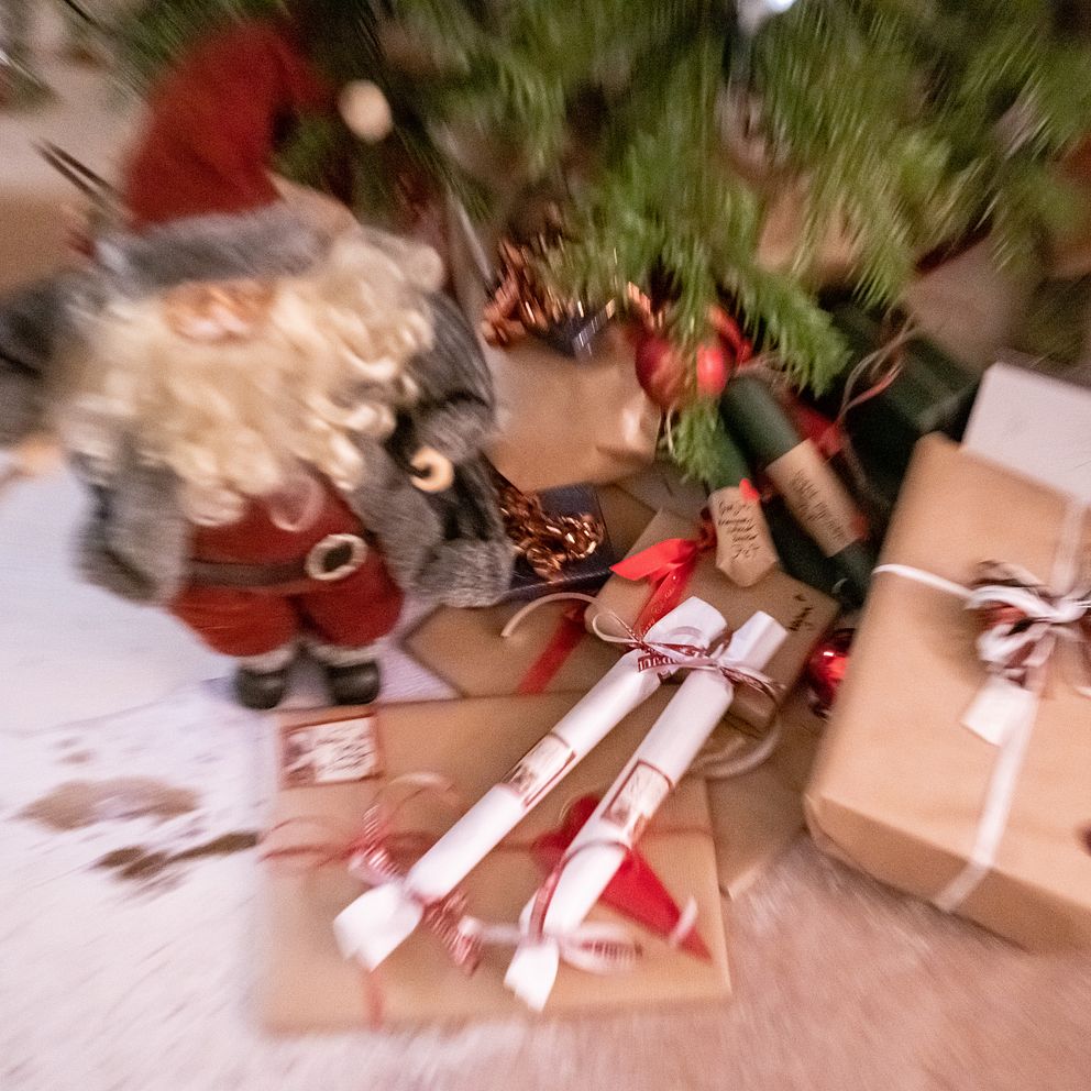 Juldekorationer med en leksakssanta, inslagna presenter och rullar med band ligger under ett julgran. Bakerst syns gröna och röda färger som utsmyckning. Bilden har en otydlig effekt, vilket ger en suddig känsla.