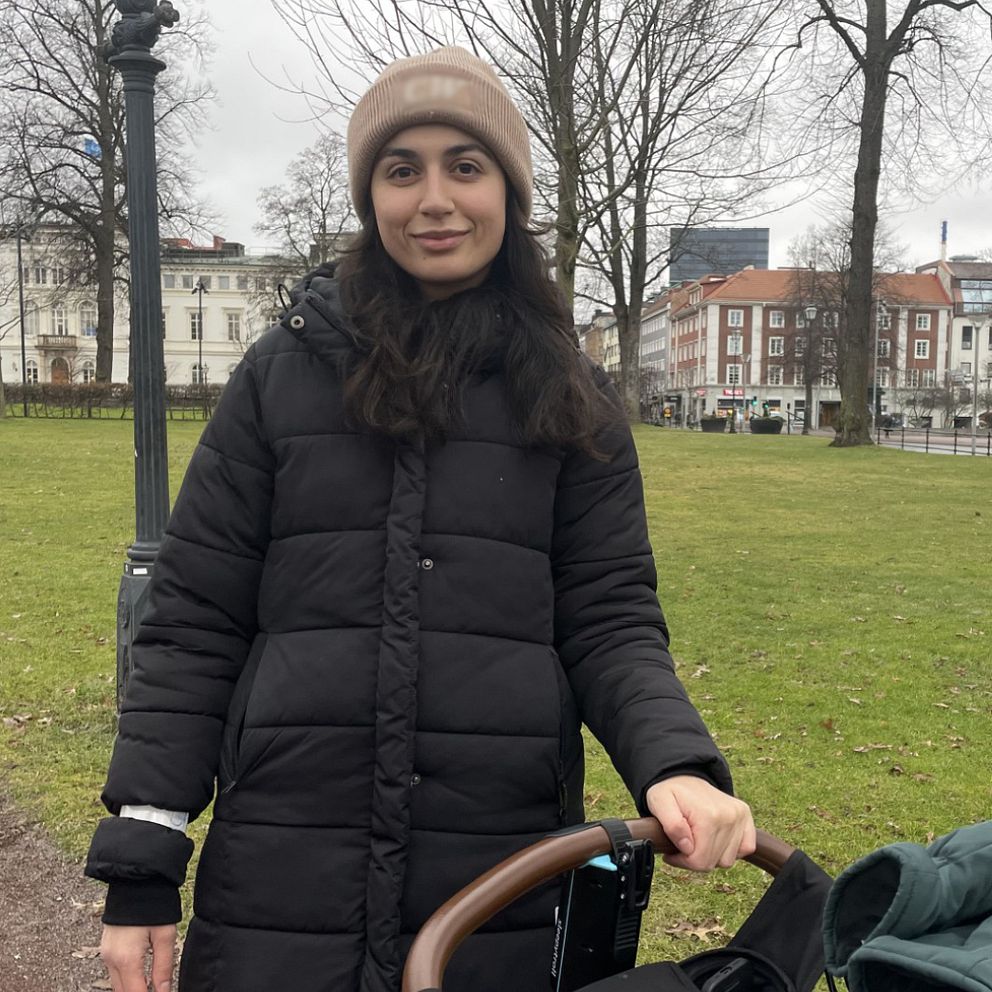 En kvinna klädd i en svart vinterjacka står i en park och håller i handtaget på en barnvagn. Det är en molnig dag, och omgivningen består av gräsytor, träd och några byggnader i bakgrunden.