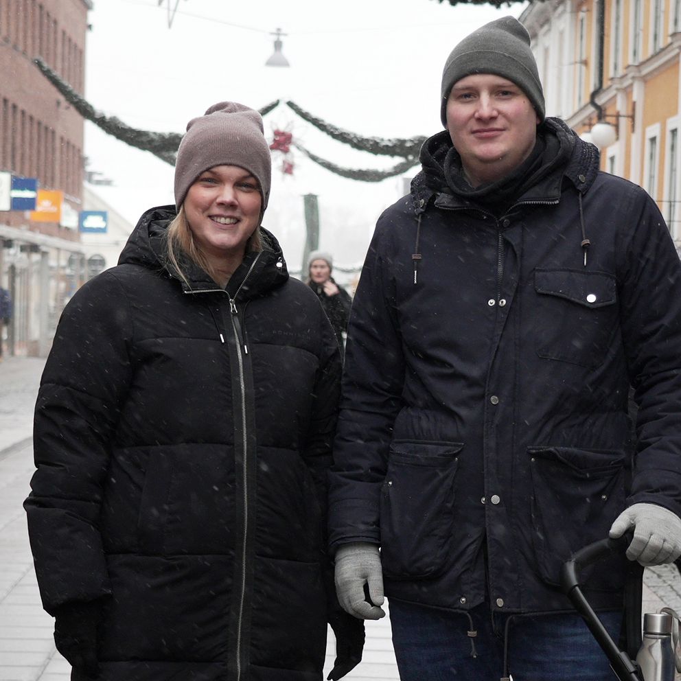 Två personer klädda i vinterjackor står på en snötäckt gata. I bakgrunden syns affärer och en uteservering. Snöfall och vinterstämning präglar bilden.