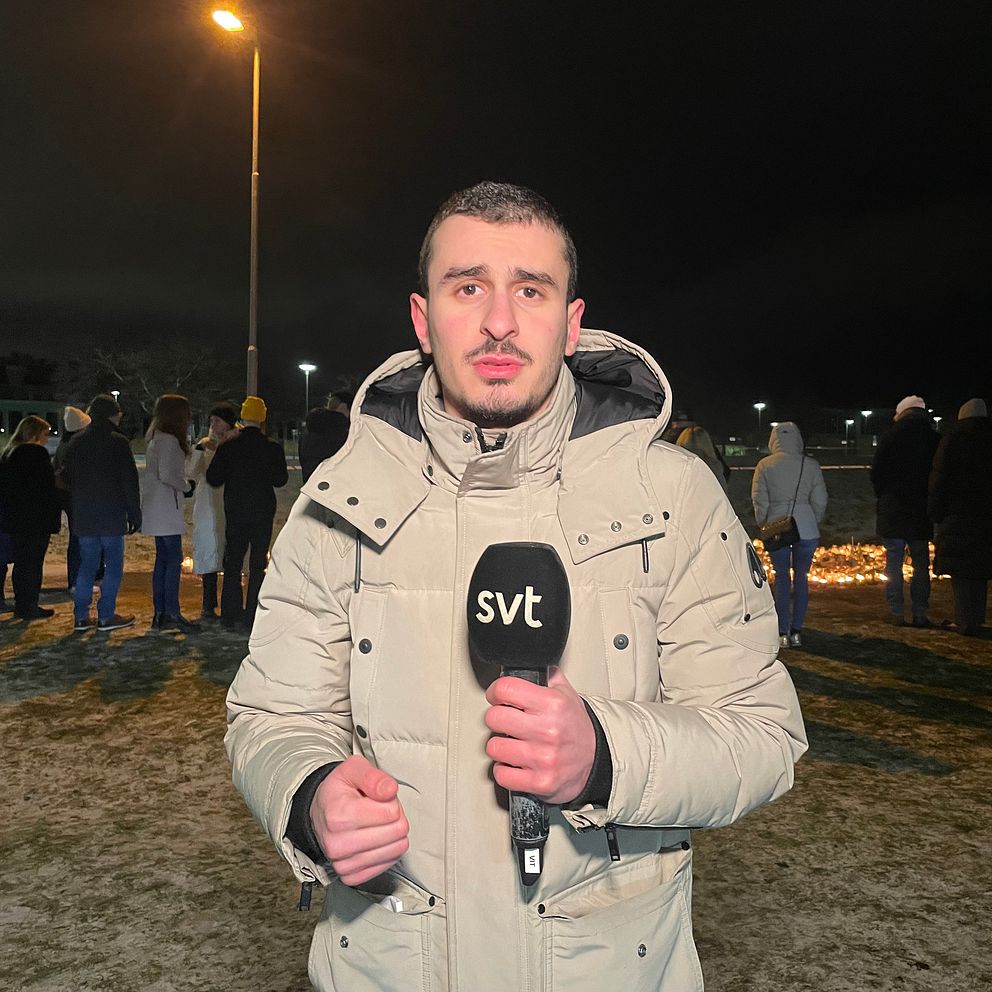 En reporter i beigefärgad jacka står utomhus, håller en SVT-mikrofon och rapporterar från en kvällshändelse med människor samlade i bakgrunden där flera ljus är tända.