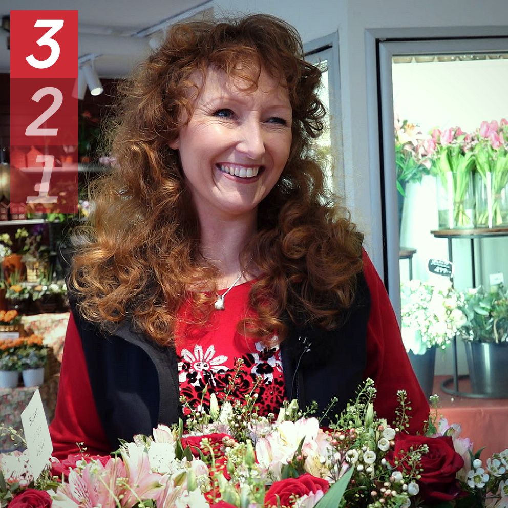 Floristen Angelica Pettersson med långt, lockigt hår står i en blomsteraffär omgiven av färgglada buketter och blommor i bakgrunden.