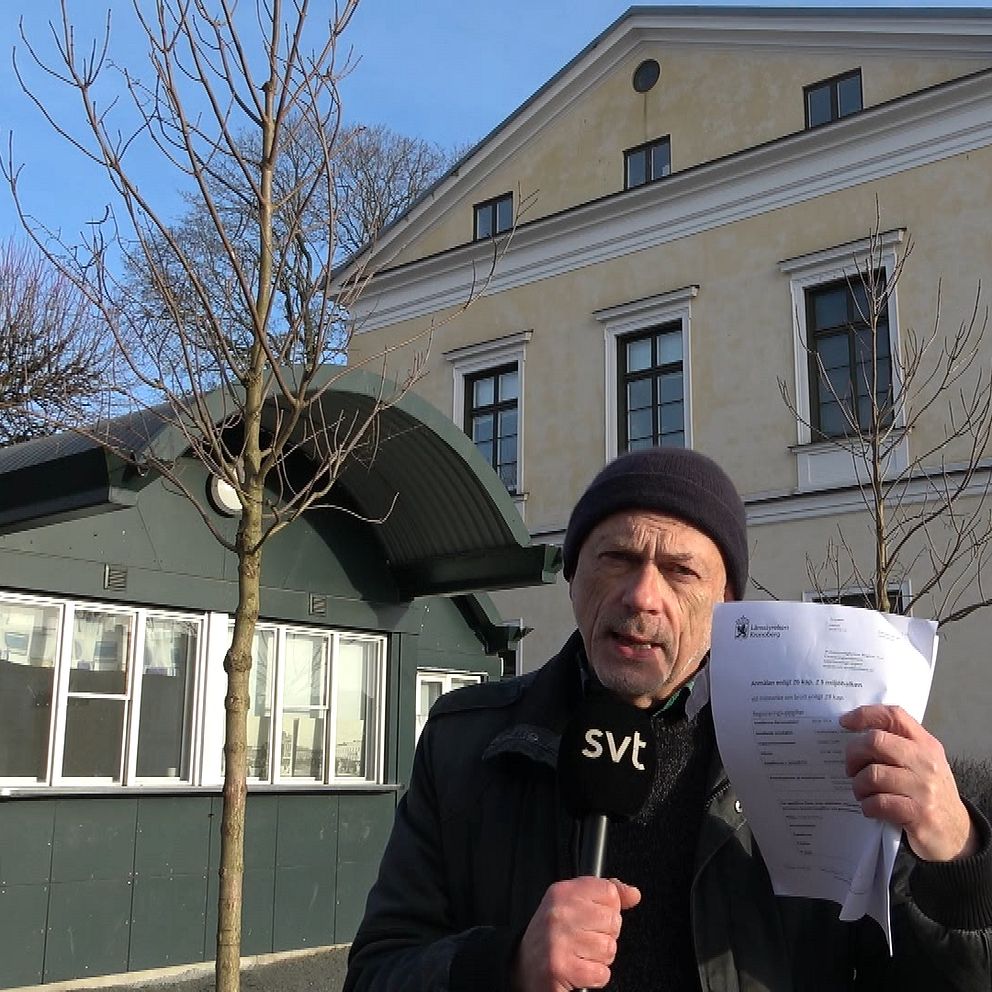En person står framför en byggnad med en trädgård och håller upp papper i ena handen. Byggnaden är i klassisk stil med ljus fasad och stora fönster. I bakgrunden syns en liten grön byggnad som kan vara ett kontor eller kiosk.
