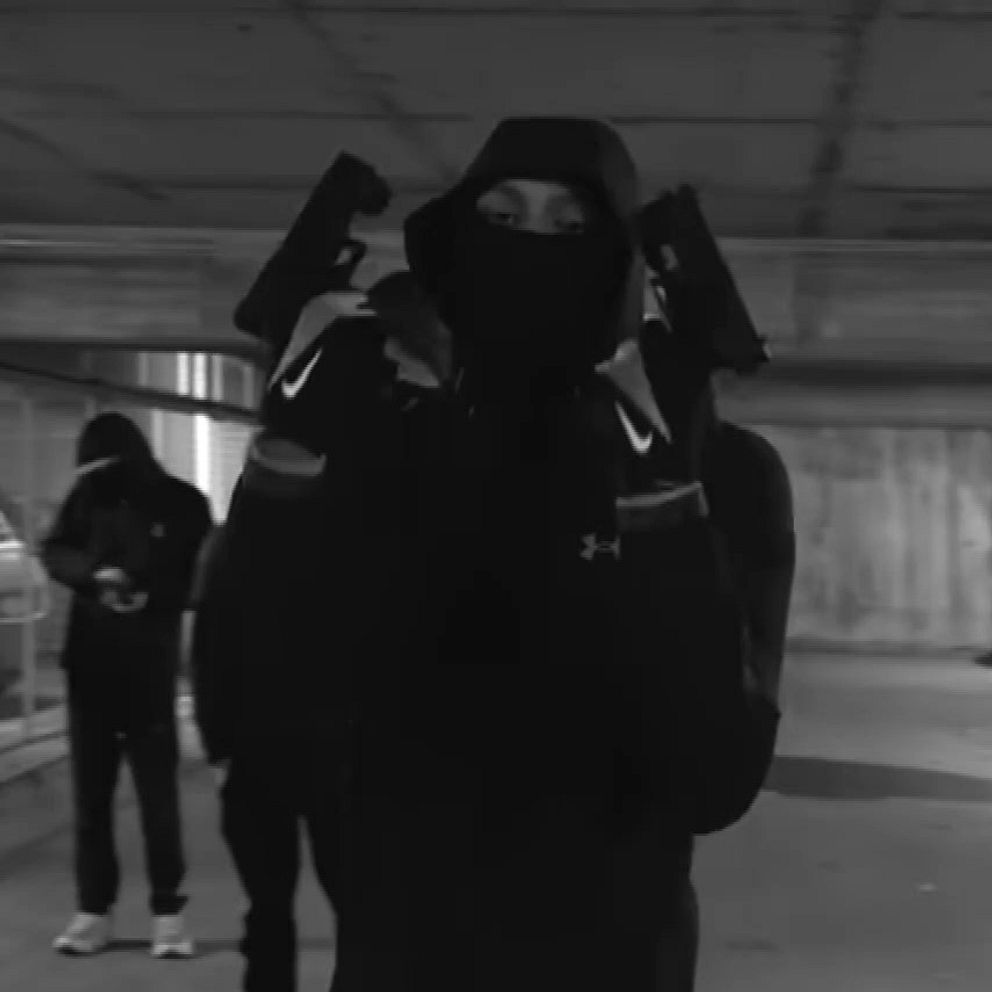 Personer i mörka kläder och masker i ett garage. Främsta personen håller två pistoler.