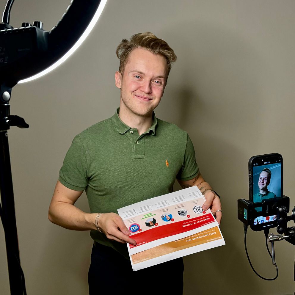 Influencern Winsent Thunholm Wikman håller i resultatet från Maktbarometern, medan en telefon monterad på ett stativ visar en ansiktsbild. Belysningen kommer från en ringlampa i bakgrunden. Rummet ser ut att vara en inomhusmiljö med moderna möbler och en köksyta synlig i bakgrunden.