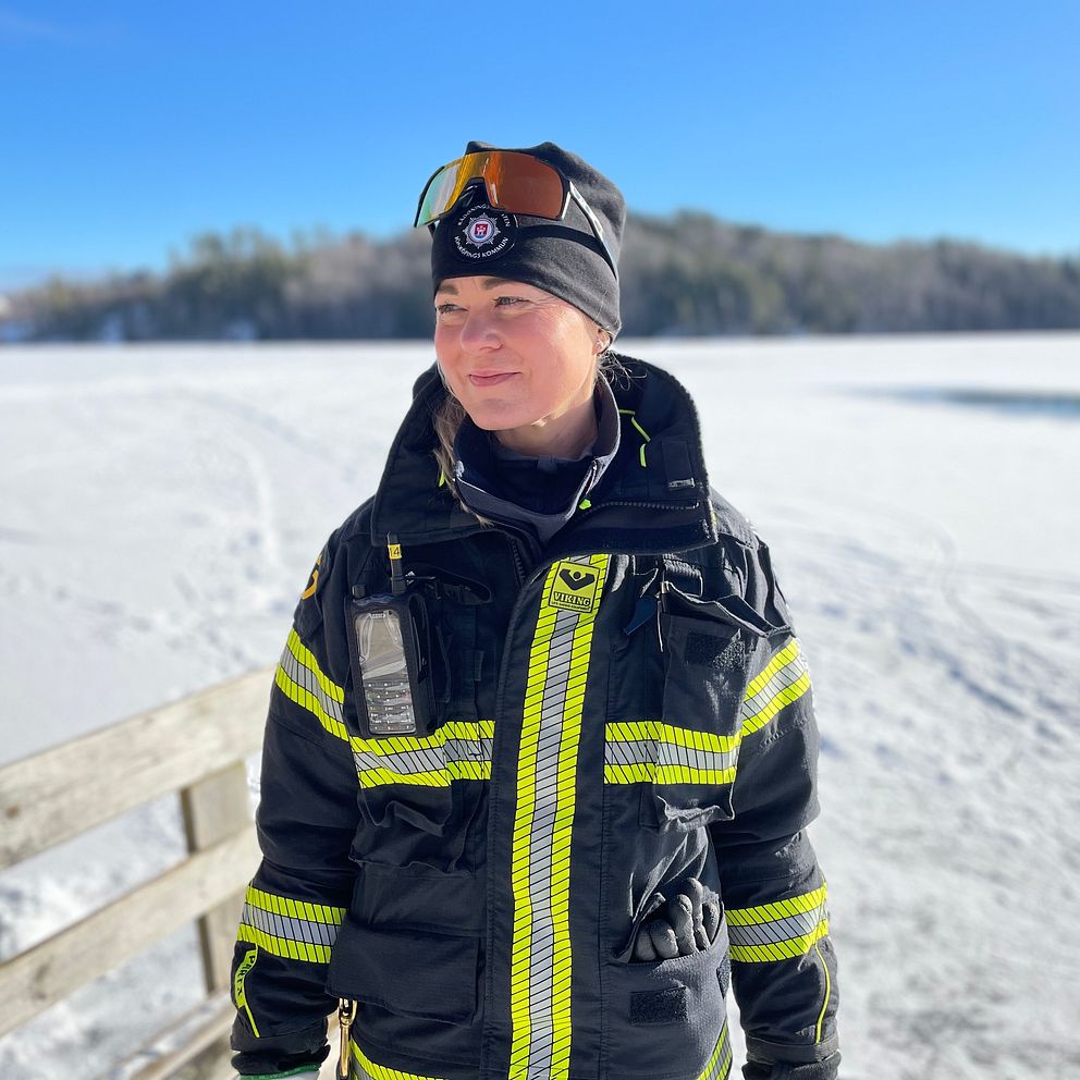 Deltidsbrandmannen Elna  i vinterkläder står vid frusen sjö i Bunn med snötäckt landskap i bakgrunden.