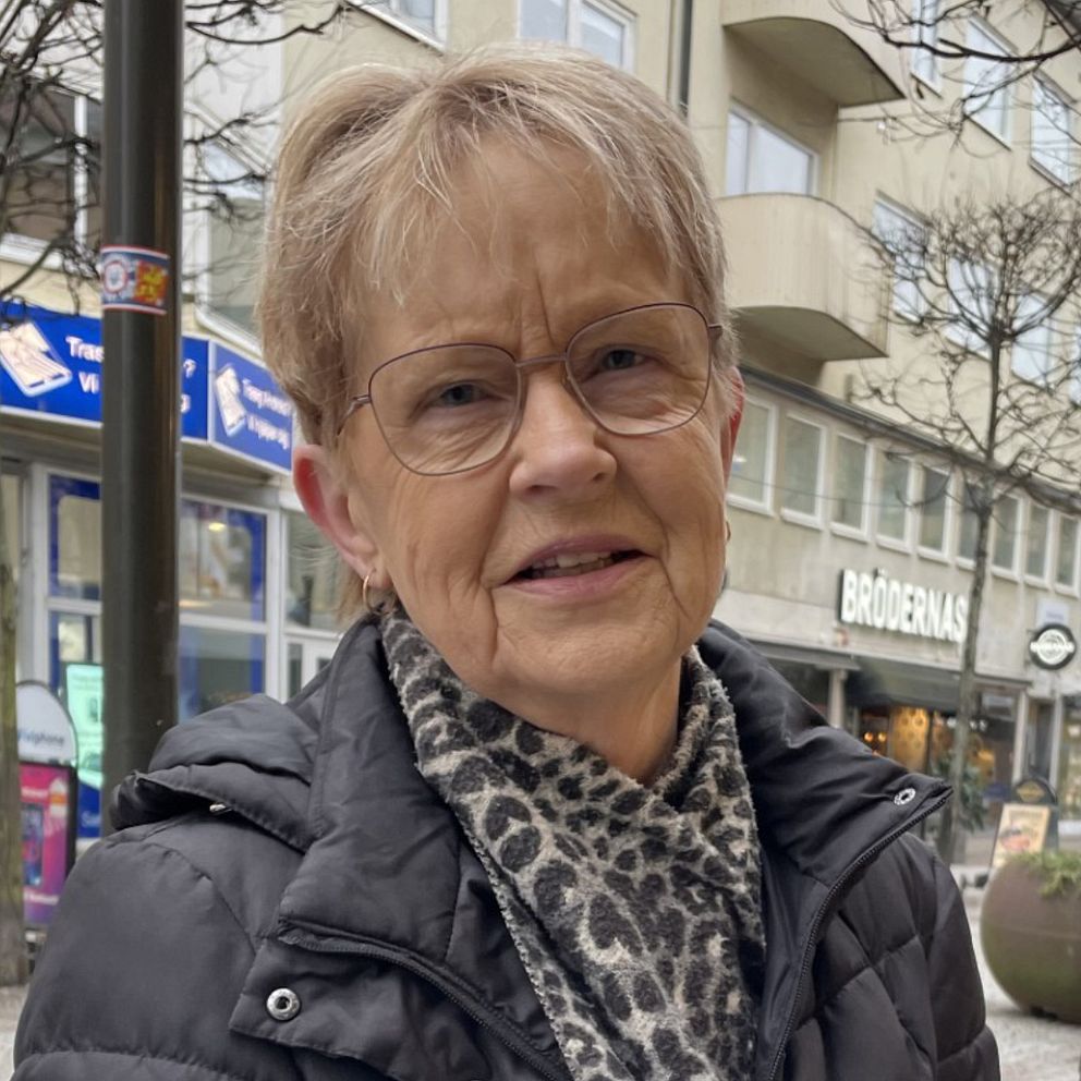 En kvinna står på en stadsgata med affärer och restauranger i bakgrunden. Träd och planteringar pryder gatan där även förbipasserande går.
