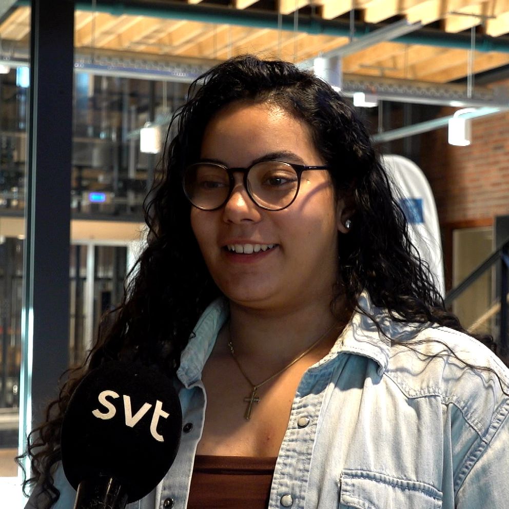 Ung kvinna intervjuas av SVT i inomhusmiljö med trädetaljer och metallstruktur.