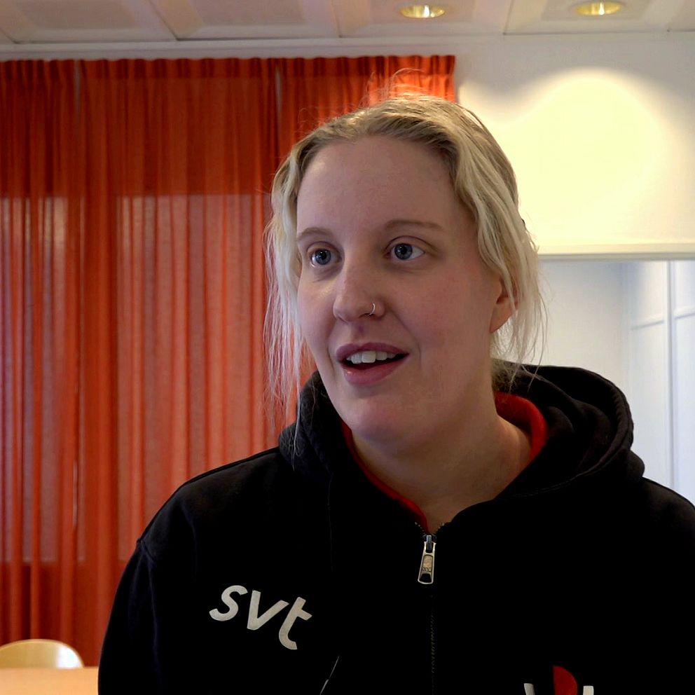 En person iklädd en mörkjacka  står i ett rum med röda gardiner och en öppen dörr i bakgrunden.