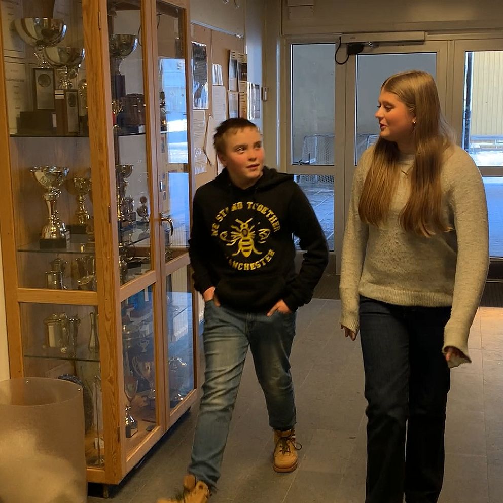 Två personer går genom en korridor i en skola eller idrottsanläggning. I bakgrunden syns en glasmonter innehållande ett flertal pokaler och priser.
