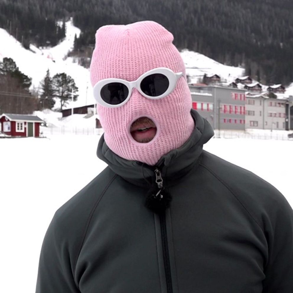 Person i mörk vinterjacka och rosa mask står utomhus på en snöklädd plats i Åre. Åre memes.