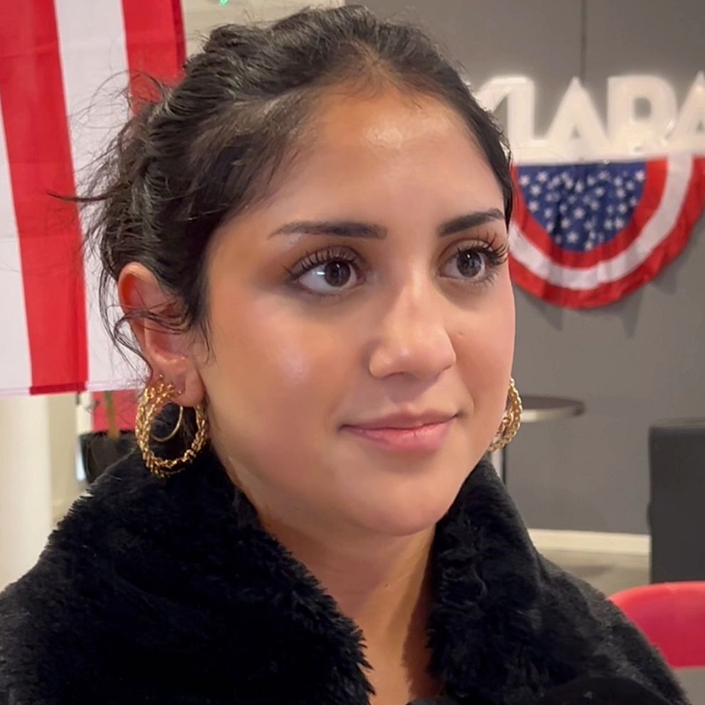 En ung kvinna står framför den amerikanska flaggan med röda, vita och blå ränder, samt en dekoration med stjärnor som hänger i bakgrunden. Hon bär stora örhängen och en svart jacka med pälsdetalj.