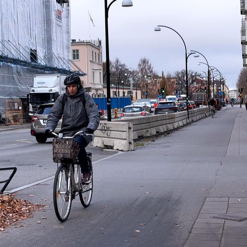En cyklist i hjälm som trampas framåt längs en bred trottoar, omgiven av en byggarbetsplats till vänster och parkerade bilar på en väg till höger. Området har ett grått väder med fallna löv längs trottoaren. Byggnaden i bakgrunden är delvis täckt av byggnadsställningar.