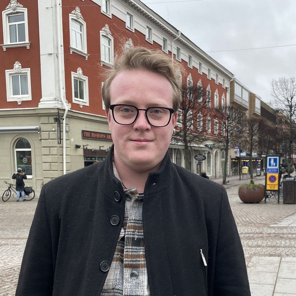En man står på en kullerstensgata. Bakom mannen syns byggnader och människor som går och cyklar.