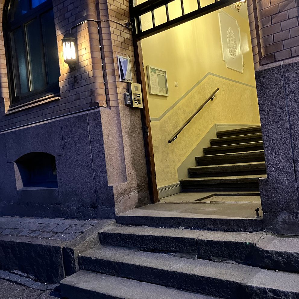 Entré till en byggnad med upplyst trapphuset på kvällstid. Trappor leder in till dörren.