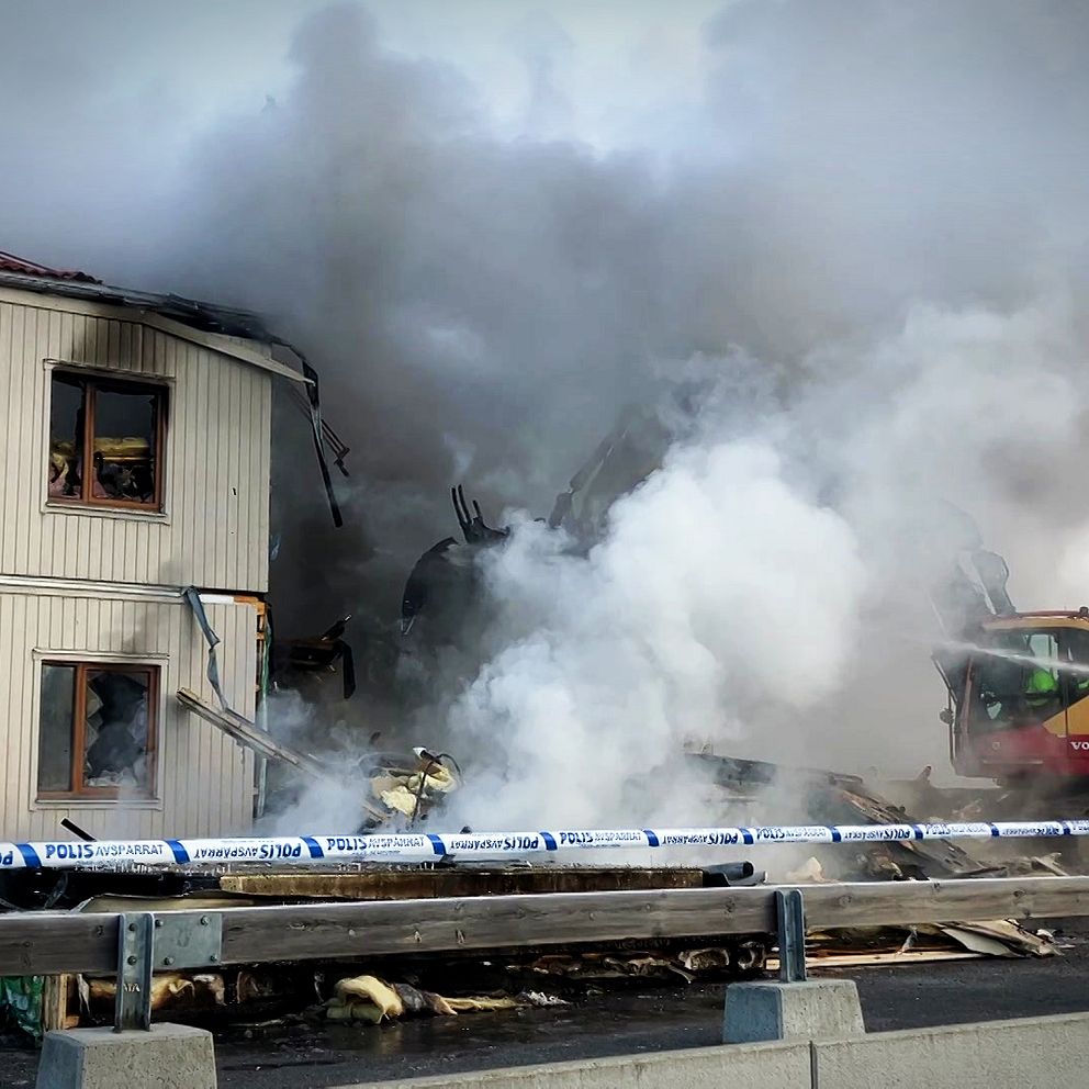Det ryker kraftigt omkring ett ljust flerfamiljshus i trä i Hölö, och räddningstjänsten sprutar vatten för att släcka branden. En grävmaskin arbetar och i förgrunden är polisens avspärrningsband.