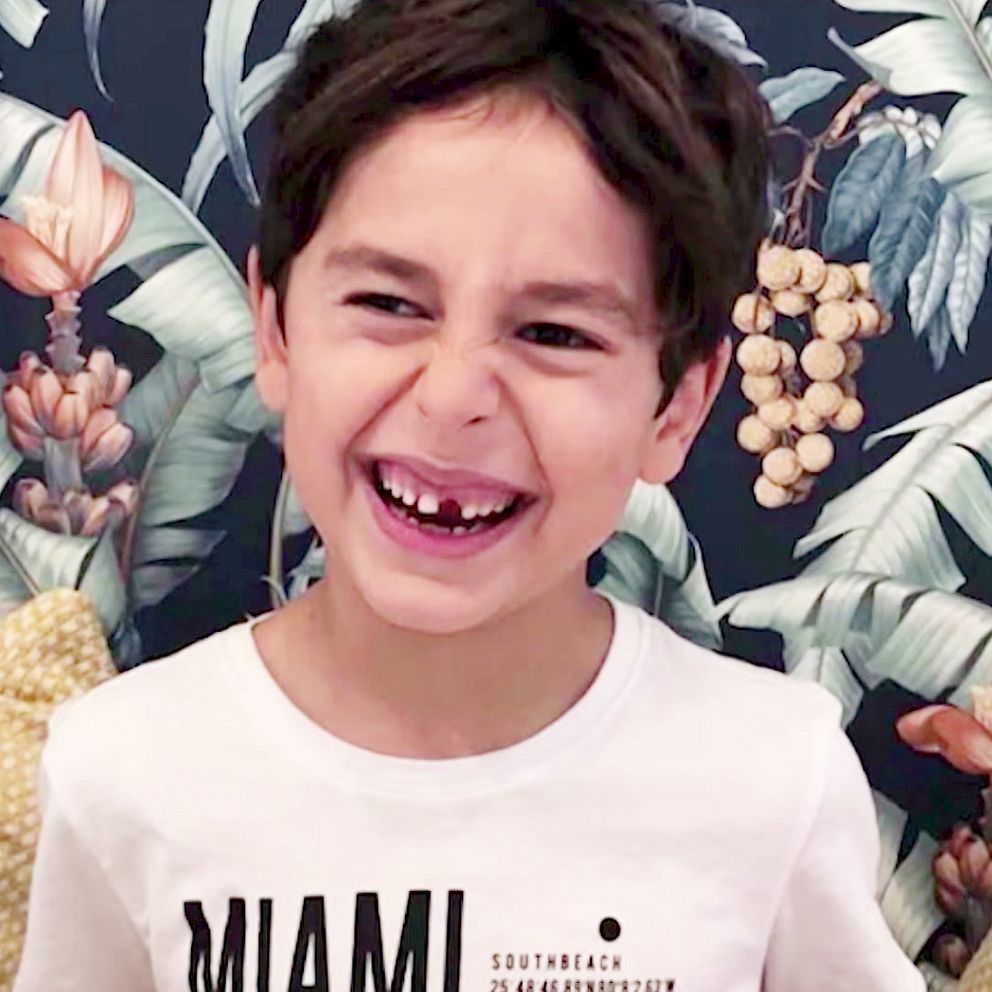 En pojke med mörkt hår , Tintin som mördades av sin pappa, sitter framför en blommig bakgrund med tropiska motiv. Han bär en vit t-shirt med texten ”MIAMI” och sitter mot kuddar med mönster.