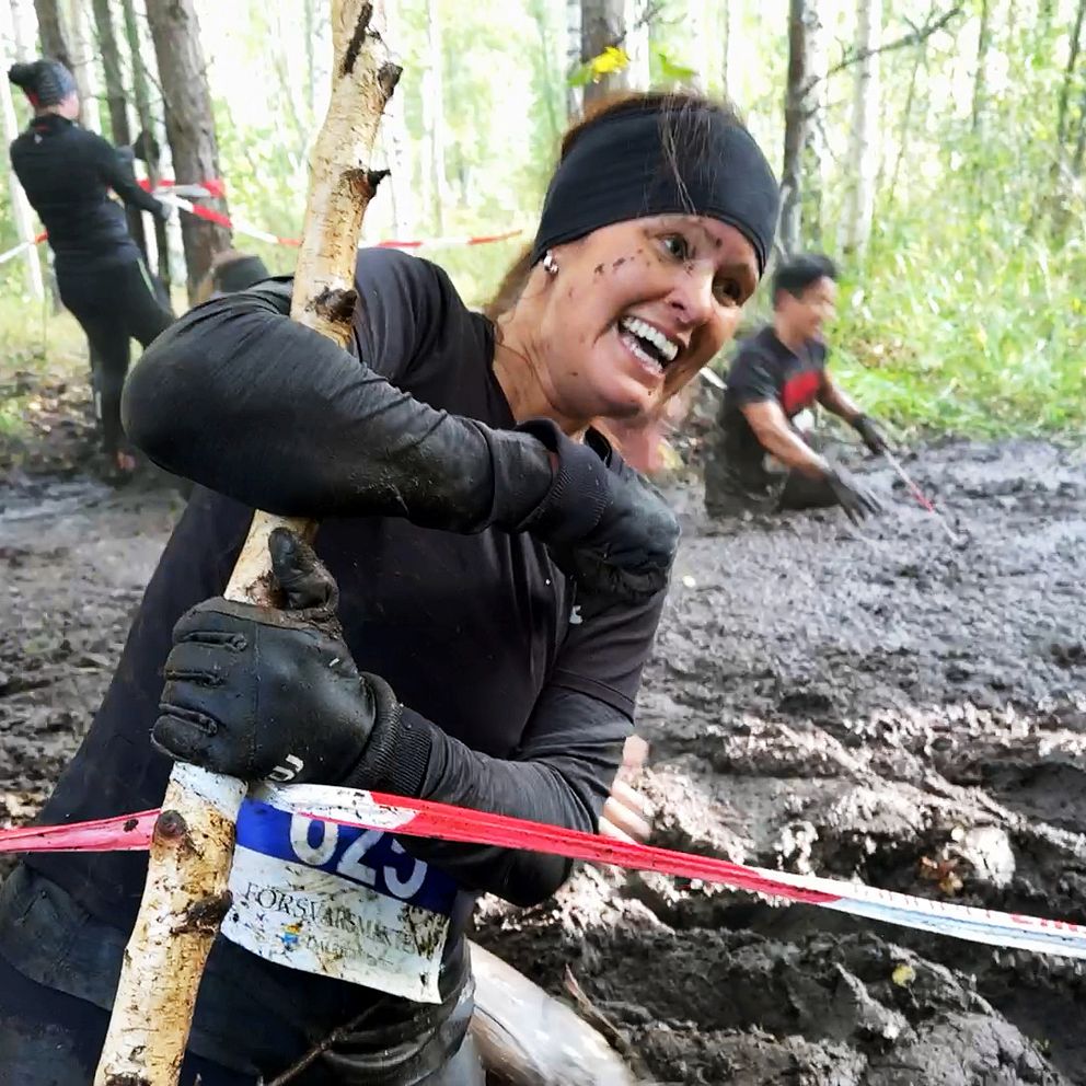 En kvinna klädd i svarta träningskläder klamrar sig fast vid en liten björk under Dala mud trail-loppet i Falun. Hon har gyttja upp till midjan, en nummerlapp på magen och ser glad men plågad ut.